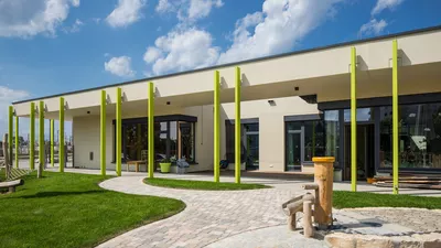 Ein einstöckiges, weißes Gebäude mit bodentiefen Fenstern und Überhang, der von grünen vertikalen Streben gehalten wird. Vor dem Haus ist eine Spielwiese mit einem gepflastertem Weg der zum Haus führt.