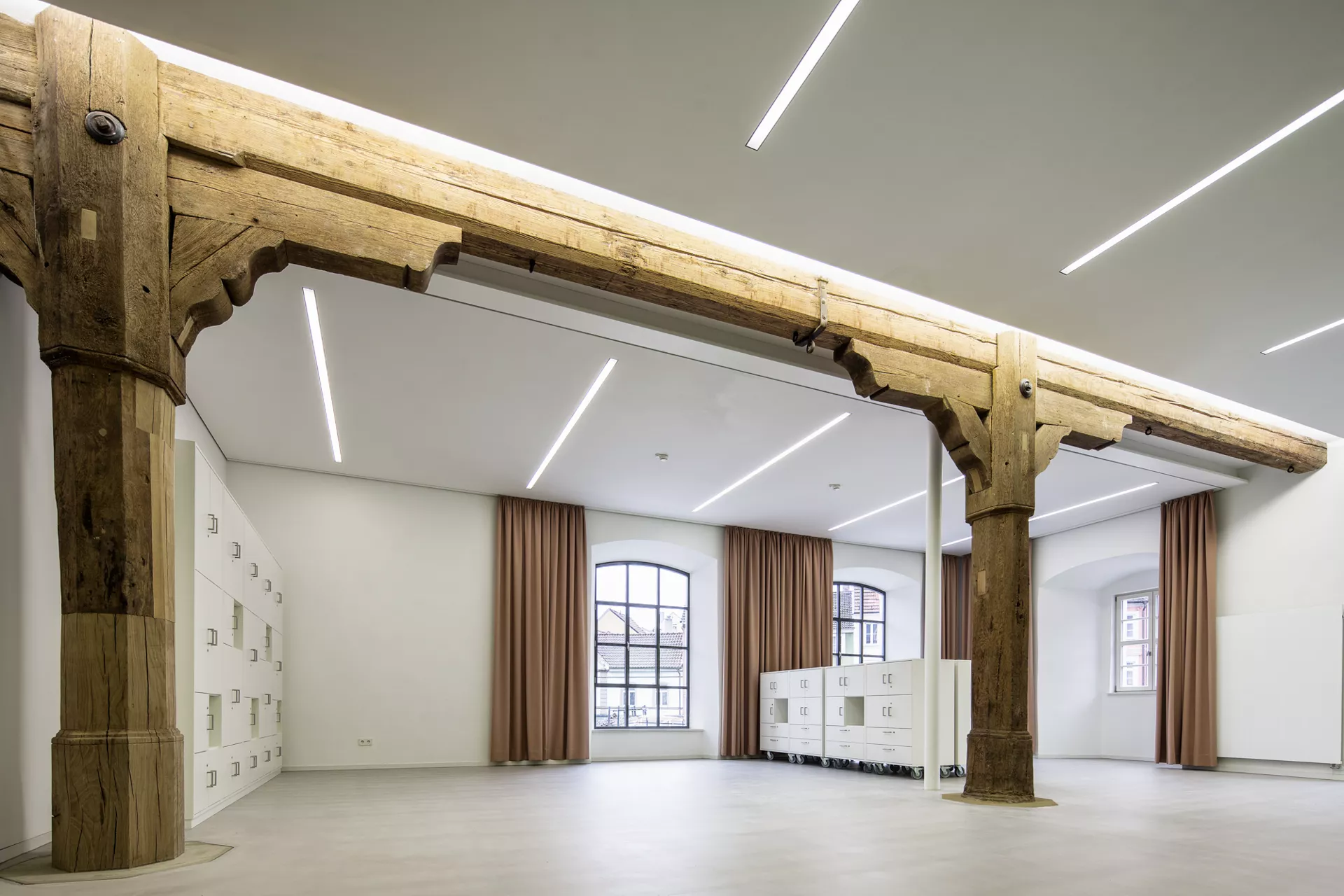 Weißer Lagerraum mit einer Schrankwand auf der linken Seite und einem niedrigen Regal mit Türen und Schubladen in der Mitte des Raums. Dekoriert wird der Raum von altertümlichen Holzträgern und -pfeilern und braunen Vorhängen an den Fenstern