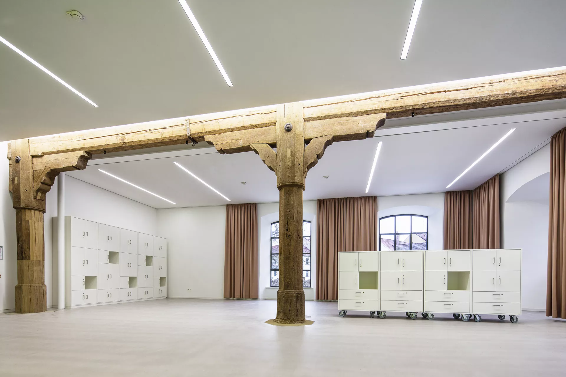 Weißer Lagerraum mit einer Schrankwand auf der linken Seite und einem niedrigen Regal mit Türen und Schubladen in der Mitte des Raums. Dekoriert wird der Raum von altertümlichen Holzträgern und -pfeilern und braunen Vorhängen an den Fenstern