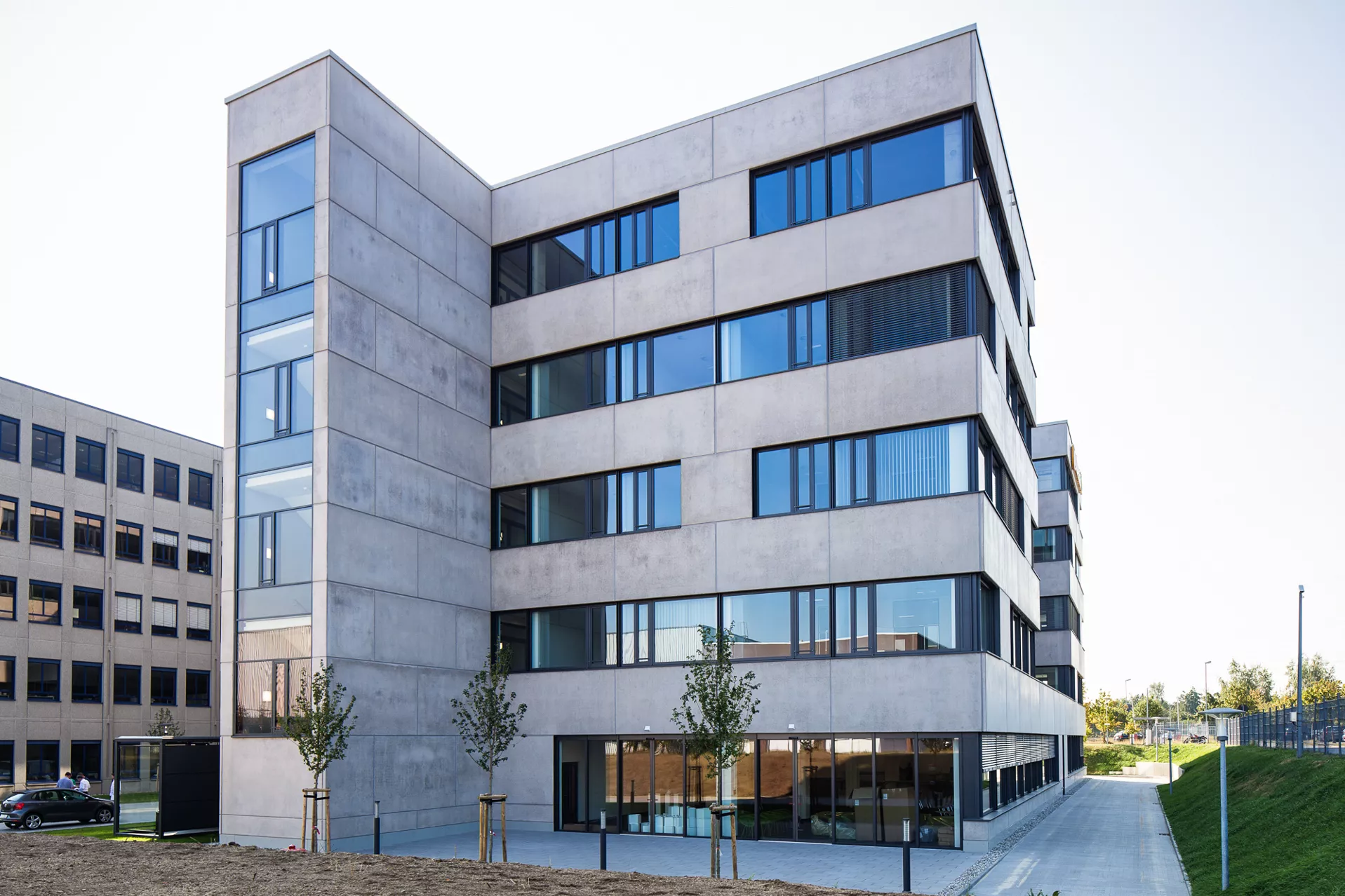 Modernes 5-stöckiges Gebäude mit polierter Betonfassade sehr breiten Fenstern. Vorne links steht das Treppenhaus hervor.