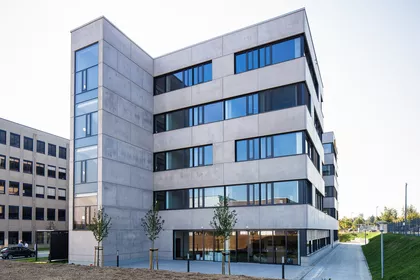 Modernes 5-stöckiges Gebäude mit polierter Betonfassade sehr breiten Fenstern. Vorne links steht das Treppenhaus hervor.