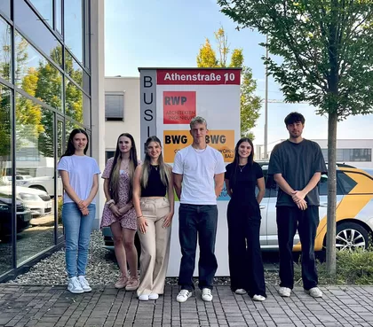 6 junge Personen auf einem gepflastertem Zugangsweg vor einem Werbeschild. Links eine Glasfassade, hinter ihnen einen quer zum Fotografen parkendes Auto und ein Baum.