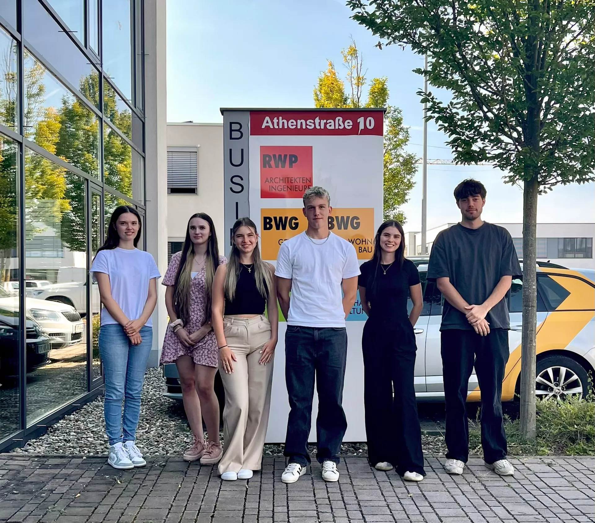 6 junge Personen auf einem gepflastertem Zugangsweg vor einem Werbeschild. Links eine Glasfassade, hinter ihnen einen quer zum Fotografen parkendes Auto und ein Baum.