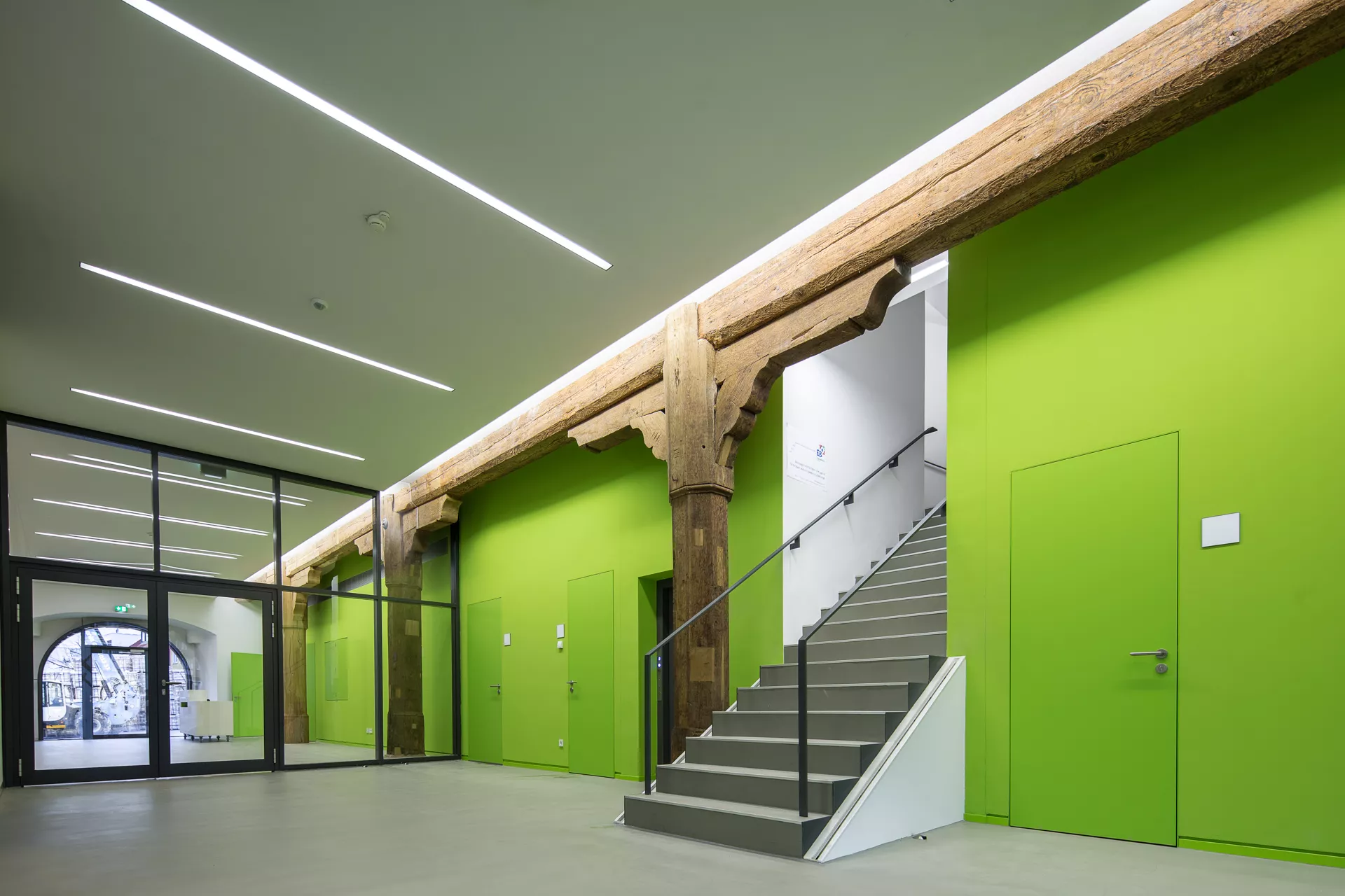 Eine grüne Wand und weiße Decke, die Decke wird getragen von einem altertümlichen Querträger und Pfeiler aus Holz. In der Mitte der Wand führt eine Treppe nach oben. Entlang der Wand befindet sich eine Glaswand mit eingelassener Tür.
