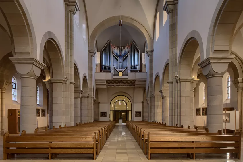 Der Hauptraum einer Kirche, fotografiert vom Alter in Richtung der Eingangstür entlang des Mittelgangs zwischen den Sitzbänken. Über der Schwelle nahe der Eingangstür befindet sich eine Orgel.