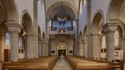 Der Hauptraum einer Kirche, fotografiert vom Alter in Richtung der Eingangstür entlang des Mittelgangs zwischen den Sitzbänken. Über der Schwelle nahe der Eingangstür befindet sich eine Orgel.
