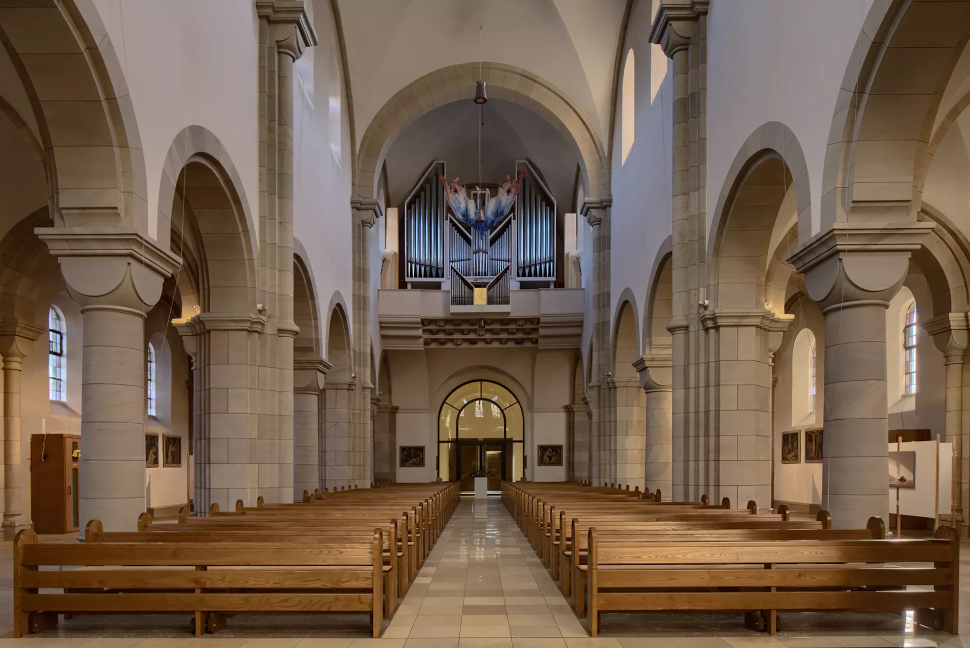 Der Hauptraum einer Kirche, fotografiert vom Alter in Richtung der Eingangstür entlang des Mittelgangs zwischen den Sitzbänken. Über der Schwelle nahe der Eingangstür befindet sich eine Orgel.