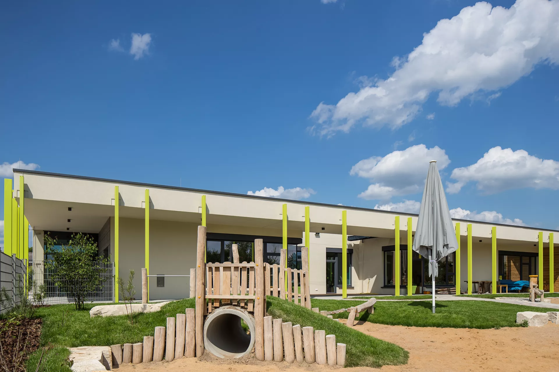 Ein einstöckiges, weißes Gebäude mit bodentiefen Fenstern und Überhang, der von grünen vertikalen Streben gehalten wird. Vor dem Haus ist ein Spielplatz mit großen Sandkasten und einer horizontalen Röhre zum durchklettern.