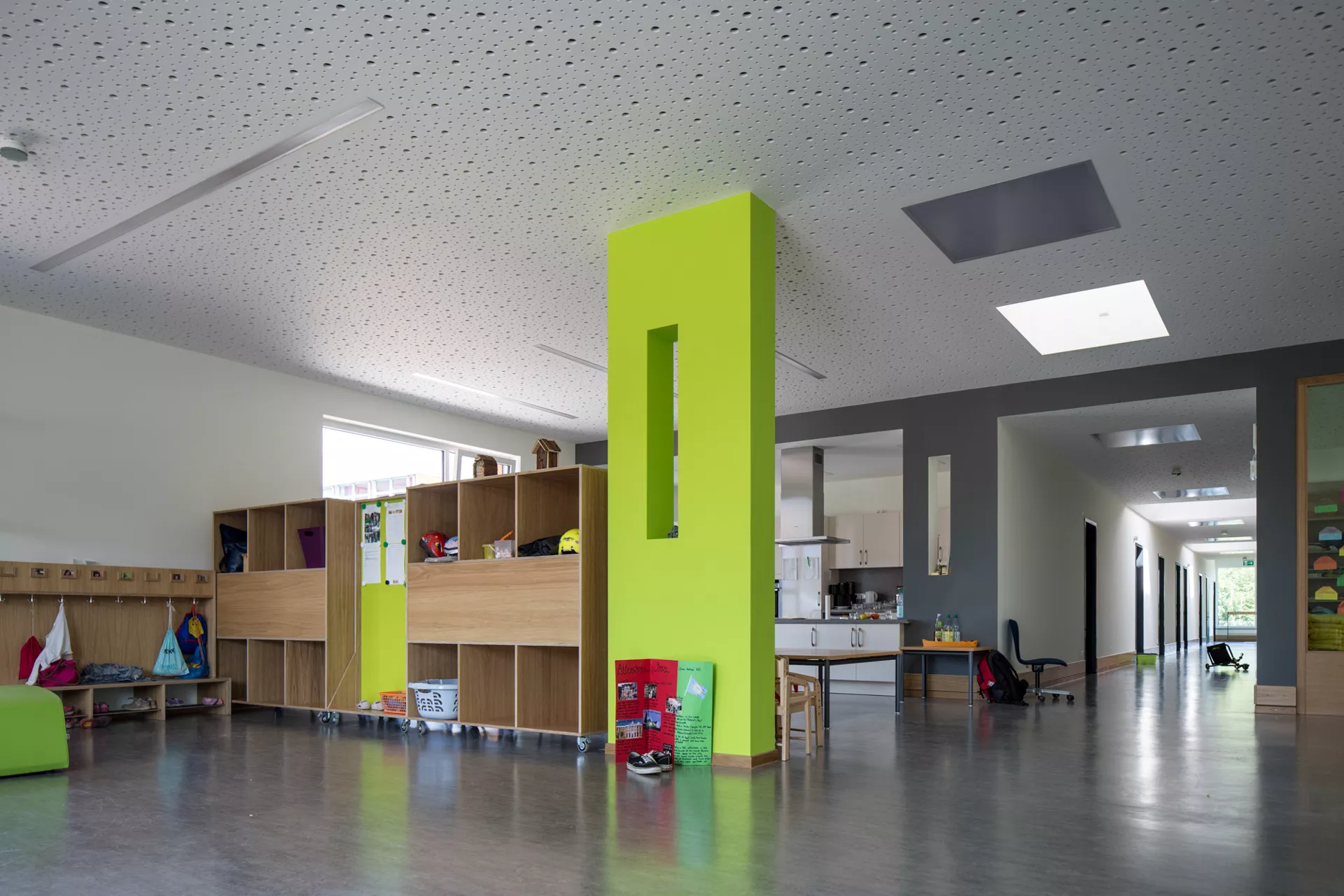 Großer Gemeinschaftsraum, grüne Säule in der Mitte. Links Garderobe mit Schuhen, Turnbeuteln und Jacken. Dahinter eine Küchezeile, rechts neben der Küche führt ein Flur tiefer ins Gebäude. 