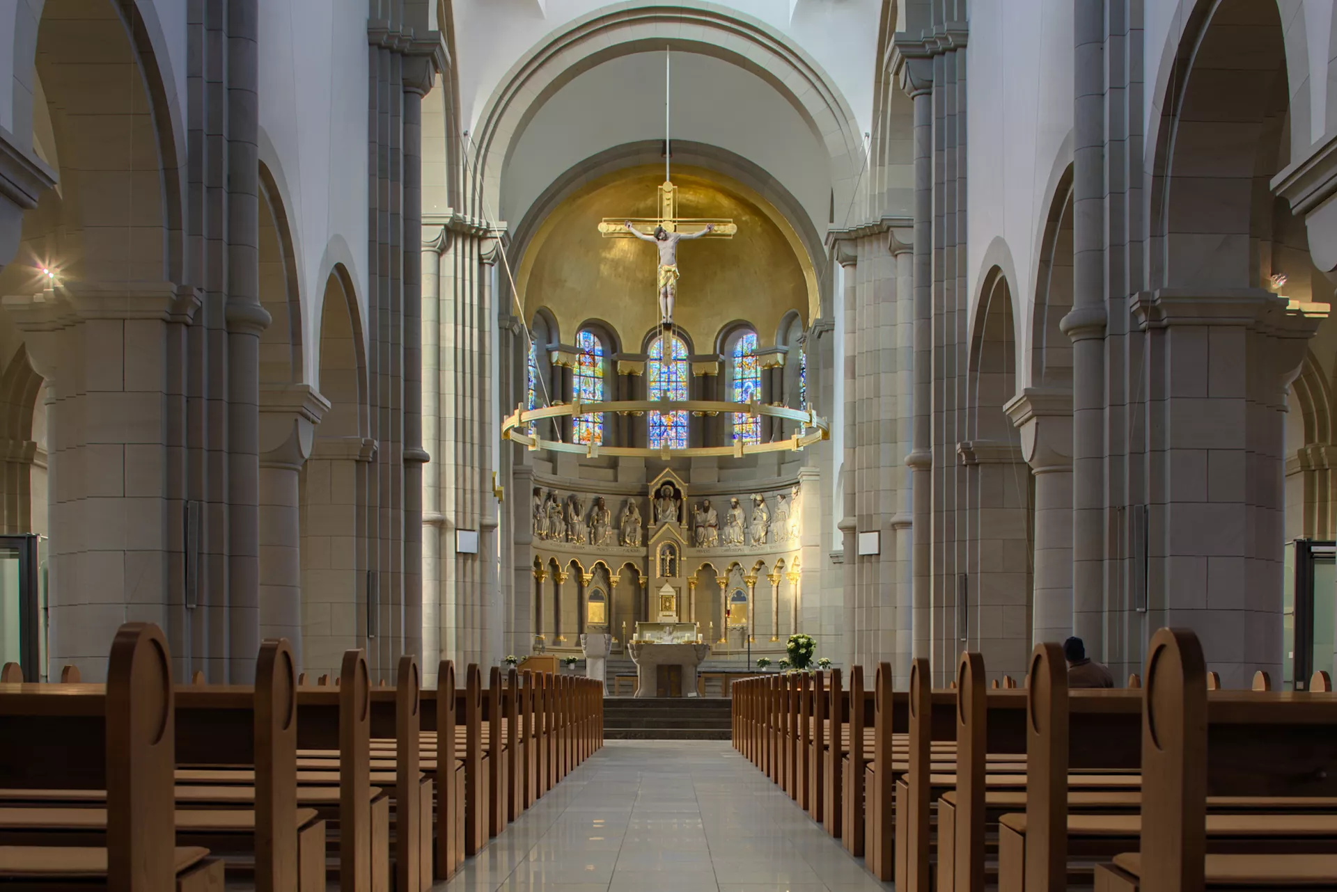 Der Hauptraum einer Kirche, fotografiert vom Eingang in Richtung des Altars entlang des Mittelgangs zwischen den Sitzbänken. Über dem Altar hängen ein runder Kerzenleuchter sowie ein großes Jesuskreuz von der hohen Decke.