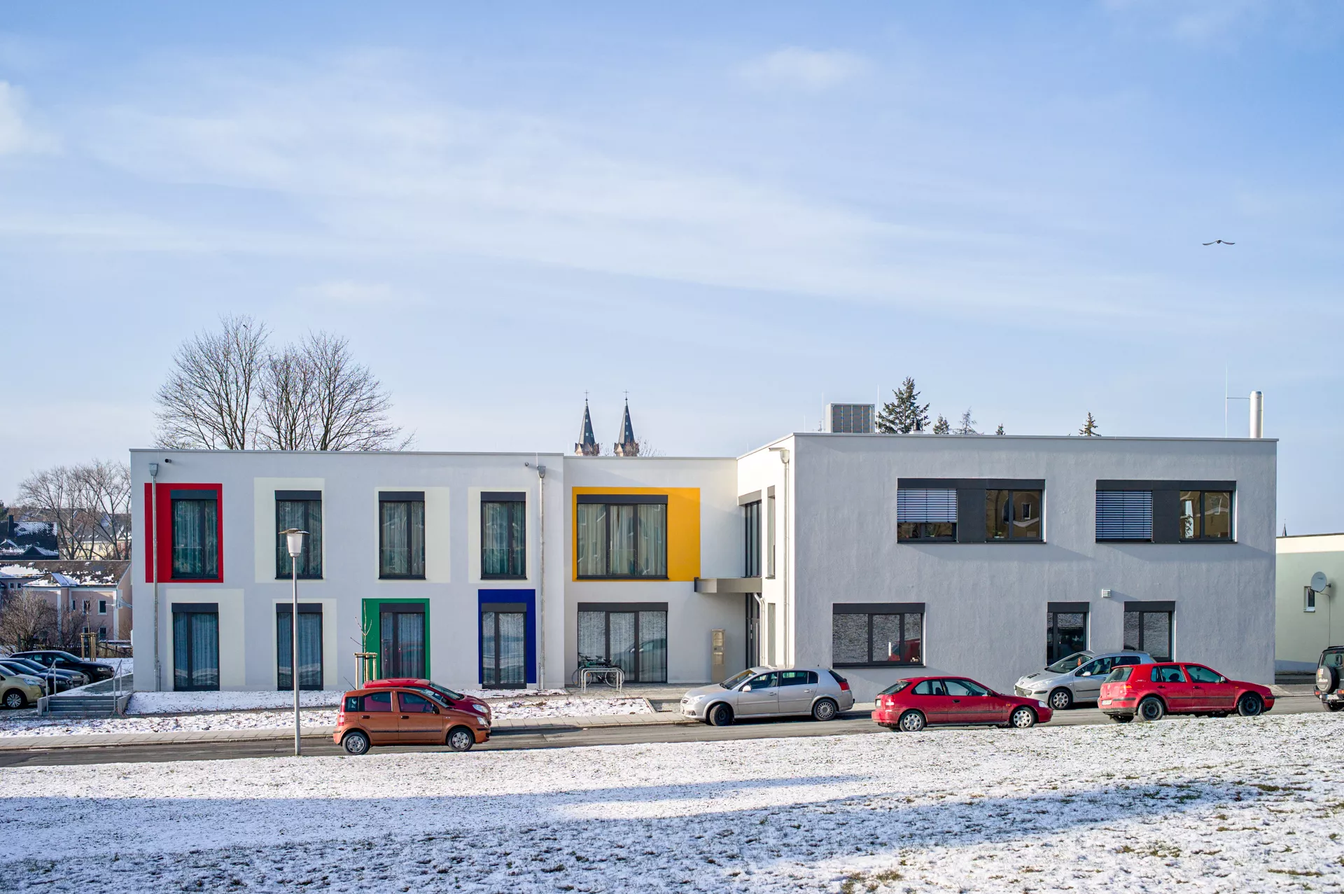 Modernes 2-stöckiges Gebäude mit bodentiefen Fenstern und Farbakzenten um manche Fenster (rot, grün, blau, gelb), schneebedeckte Wiese im Vordergrund