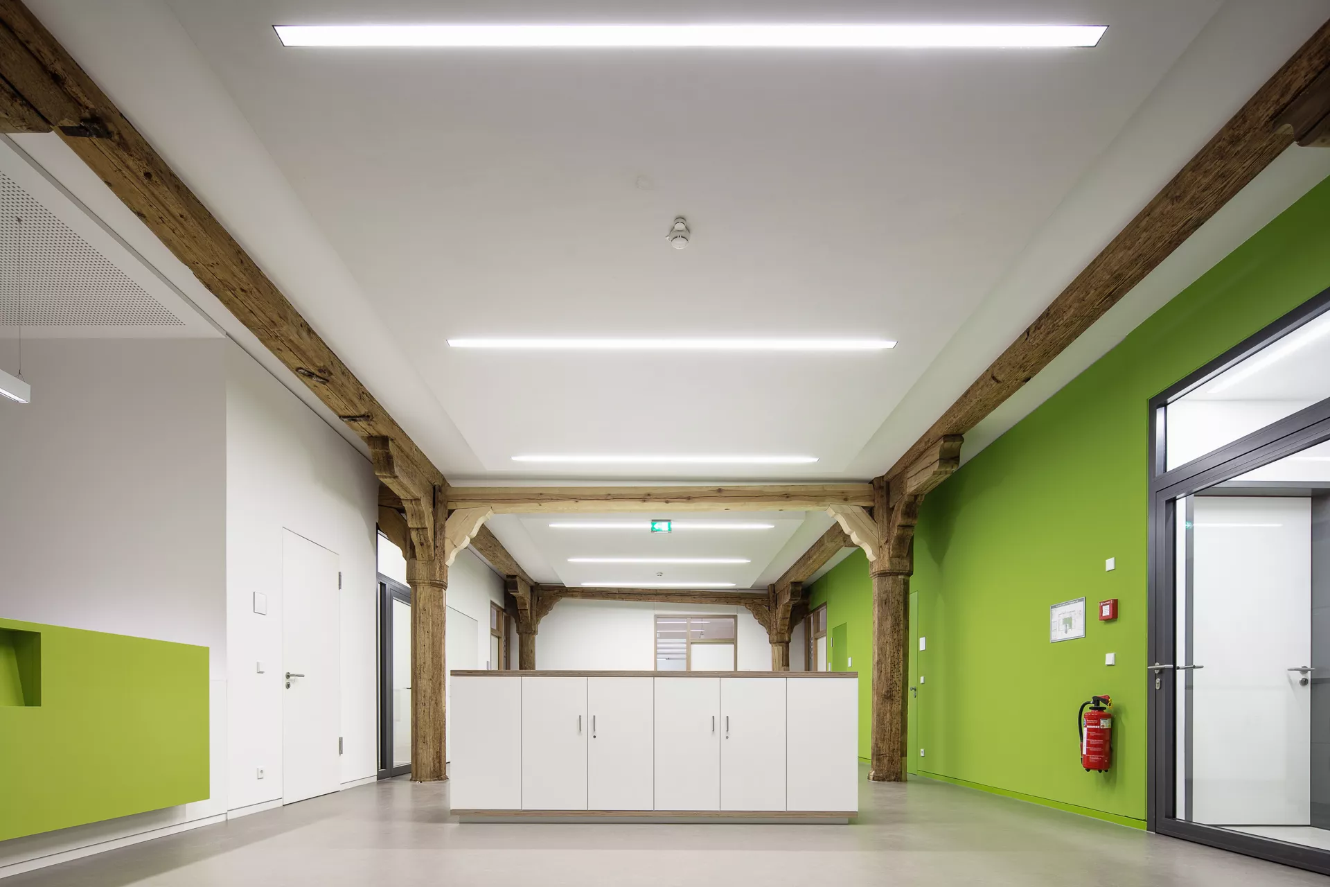 Frontansicht eines hüfthohen ca. 2,50m breiten Schrankes in der Mitte des Raumes. Links Theke mit grüner Front, rechts grün gestrichene Wand mit Durchgangstür (Glas) und Feuerlöscher an der Wand. Raum inkorporiert bestehende Holzträger und -pfeiler.