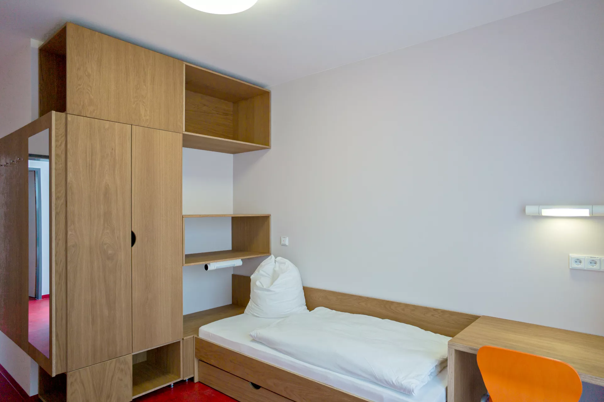 Ein einfaches Zimmer mit Bett, kleinem Schreibtisch mit Stuhl und Schrank mit Regal am Kopfende des Bettes