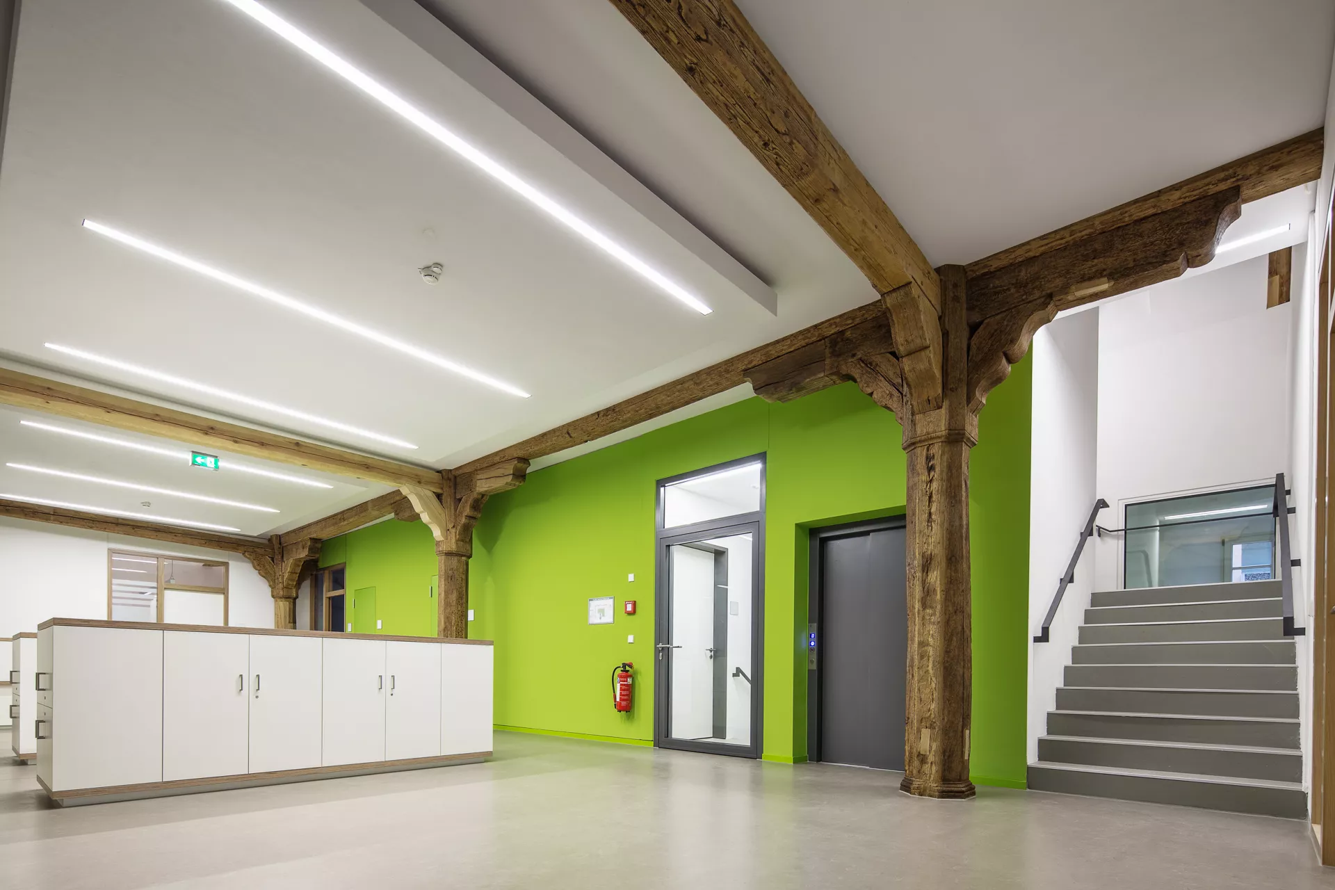 Der weiße Raum mit einer grünen Wand inkorporiert die bestehenden Holzträger und -pfeiler. In der Mitte des Raumes befinden sich hüfthohe Schränke. An der gegenüberliegenden grünen Wand führt eine Treppe nach oben und eine Glastür zur Treppe nach unten, dazwischen befinden sich Aufzugtüren.