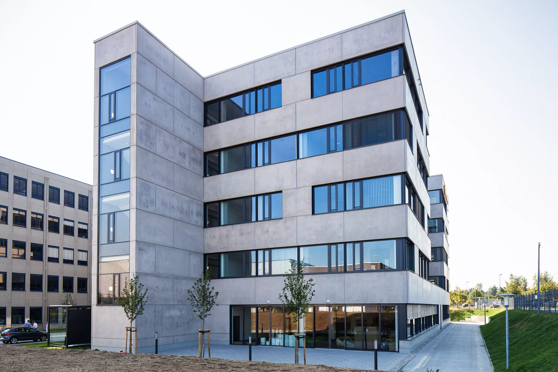 Modernes 5-stöckiges Gebäude mit polierter Betonfassade sehr breiten Fenstern. Vorne links steht das Treppenhaus hervor.