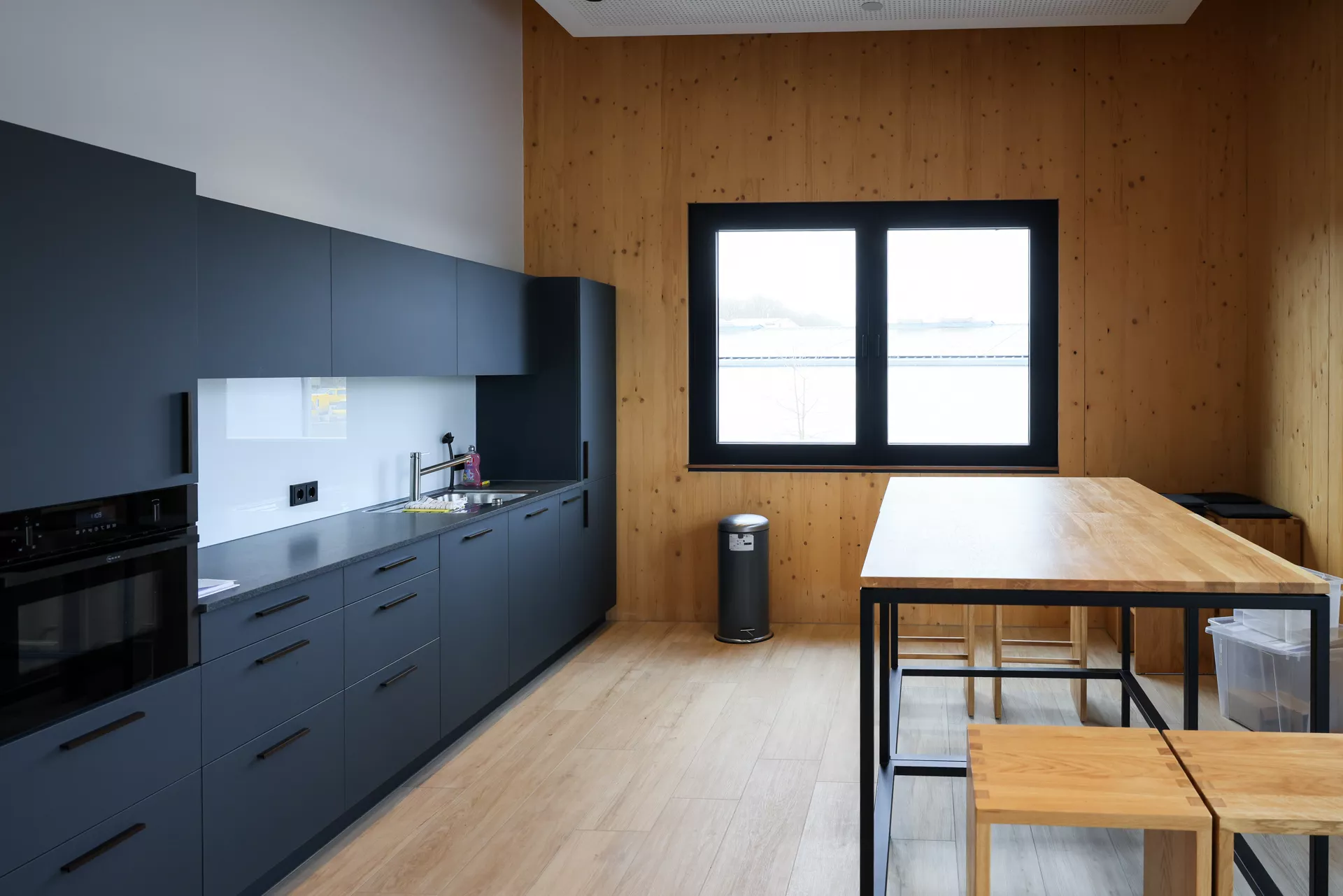 Große schwarze Küchenzeile links an der Wand und rechts davon ein Tisch mit Hochstühlen sowie ein Fenster an der hinteren Wand.