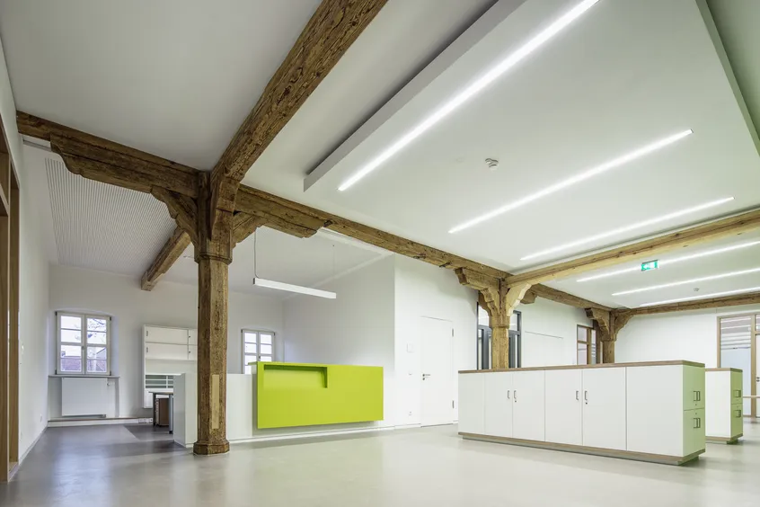 Weißer Empfangsraum mit inkorporierten Holzträgern und -pfeilern. Links befindet sich ein Arbeitsplatz mit kleiner Empfangtheke mit grüner Vorderseite, in der Mitte des Raums befinden sich hüfthohe Schränke.