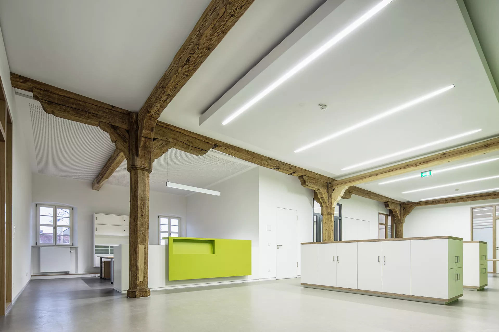 Weißer Empfangsraum mit inkorporierten Holzträgern und -pfeilern. Links befindet sich ein Arbeitsplatz mit kleiner Empfangtheke mit grüner Vorderseite, in der Mitte des Raums befinden sich hüfthohe Schränke.