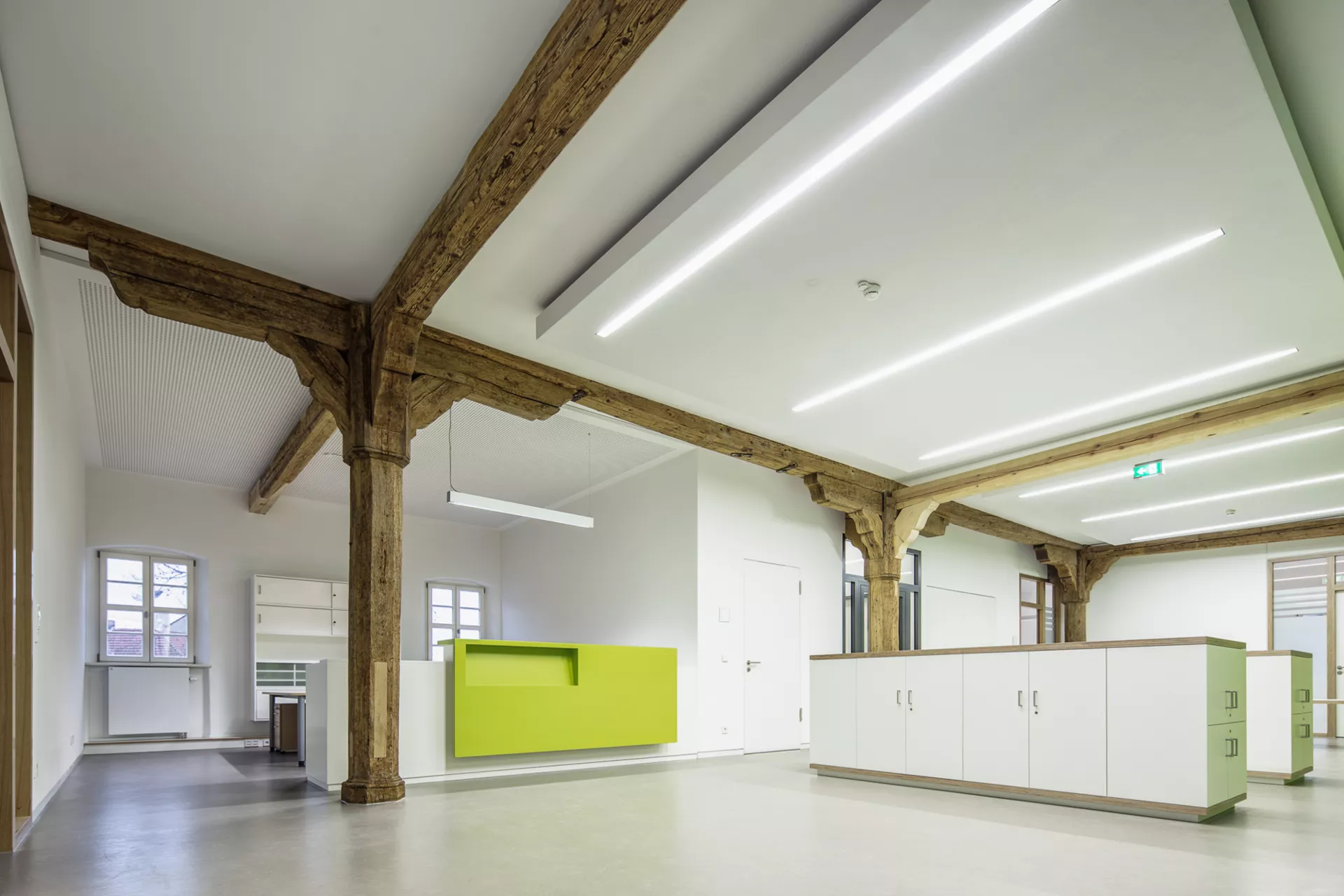 Weißer Empfangsraum mit inkorporierten Holzträgern und -pfeilern. Links befindet sich ein Arbeitsplatz mit kleiner Empfangtheke mit grüner Vorderseite, in der Mitte des Raums befinden sich hüfthohe Schränke.